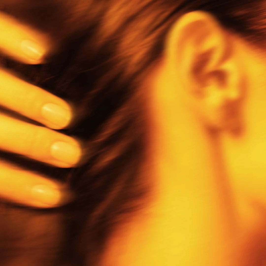 Detailaufnahme in warmen Gelb- und Orangetönen, bei der eine Hand mit gepflegten Fingernägeln durch langes Haar nahe am Ohr streicht. Das Bild ist leicht verschwommen.
