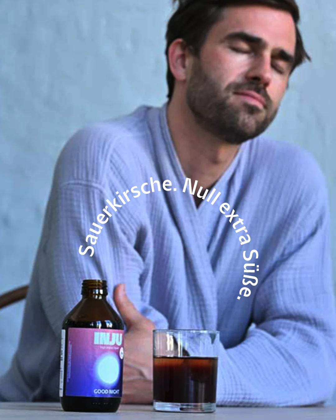 Ein Mann im blauen Bademantel sitzt mit geschlossenen Augen vor einer Flasche Good Night und einem Glas der dunkelvioletten Flüssigkeit. Text-Bogen: "Sauerkirsche. Null extra Süße."
