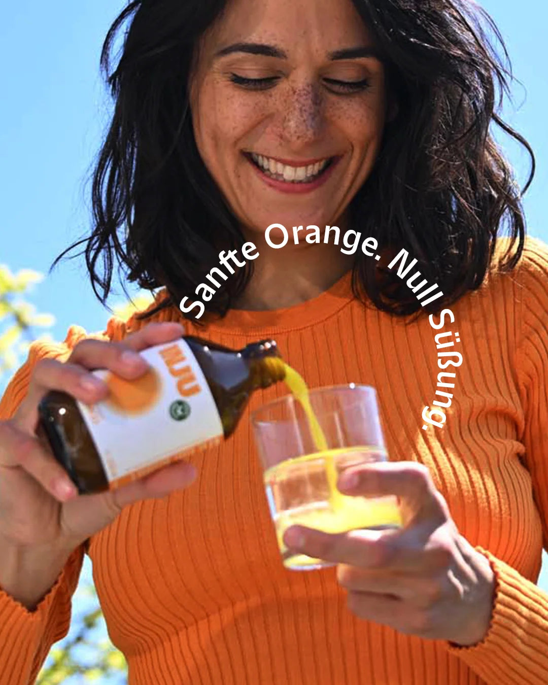 Eine lachende Frau in einem orangefarbenen Oberteil gießt die gelbe Vitamin-D3-Essenz in ein Glas Wasser. Text-Bogen: „Sanfte Orange. Null Süßung.“