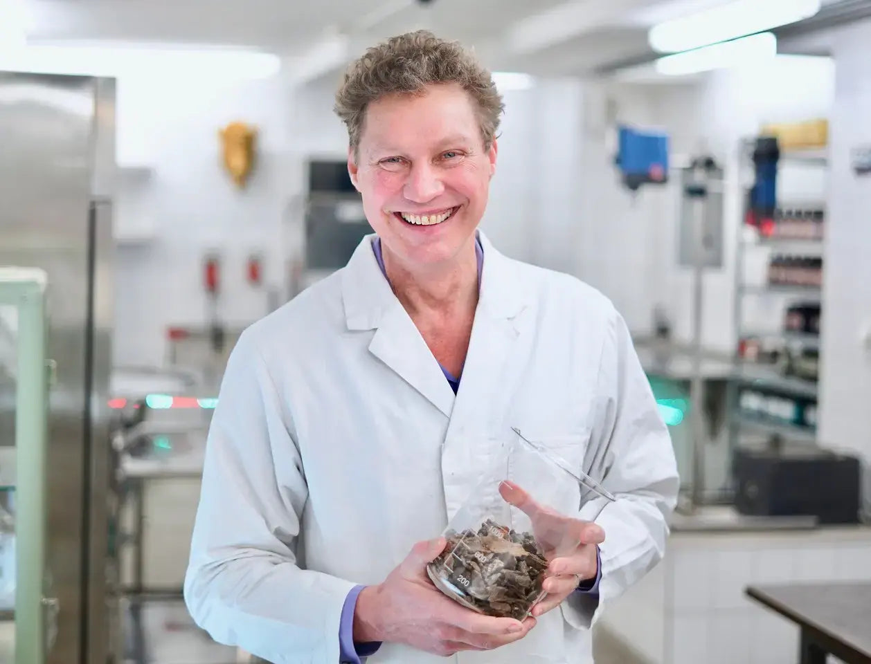 Mitgründer und Lebensmitteltechnologe Peter Kowalsky steht weißen Laborkittel in einer modernen Laborumgebung und hält ein großes Becherglas mit getrockneten Kräutern und Wurzeln in den Händen.