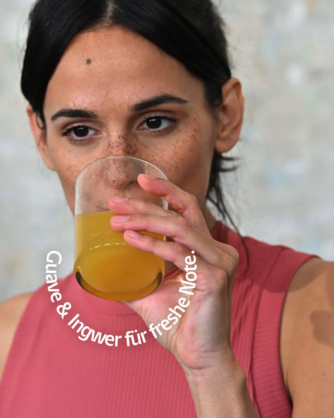 Eine Frau trinkt INJU FOCUS aus einem Wasserglas, gefüllt mit der orange-geleben Flüssigkeit. Text-Bogen: „Guave & Ingwer für freshe Note.“
