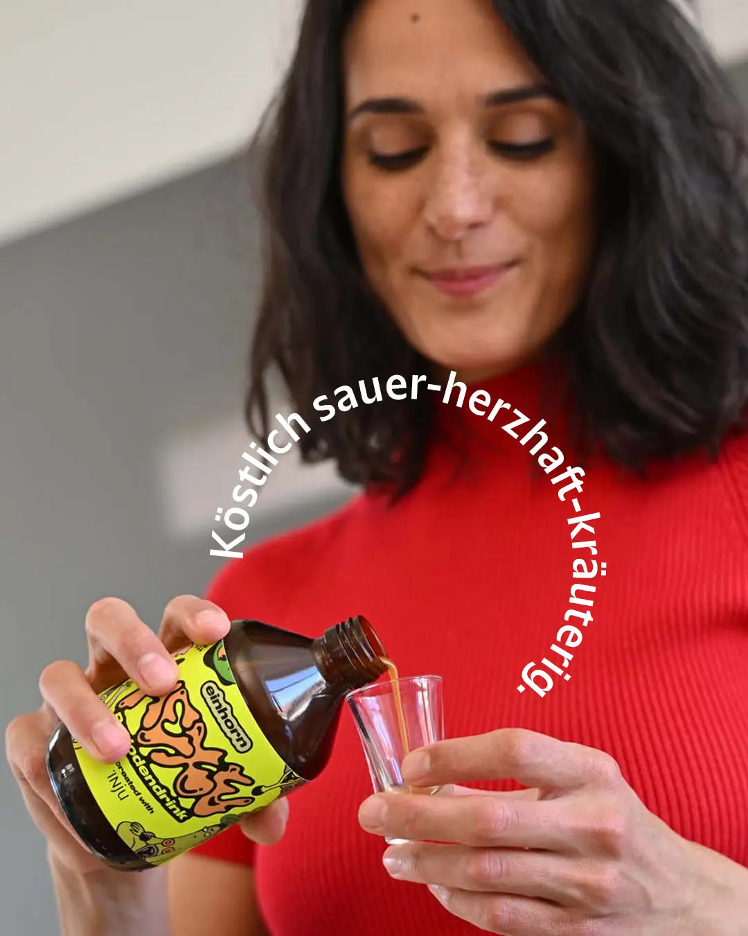 Eine Frau schenkt sich HEXE aus der gelben Flasche in ein Shotglas ein. Text-Overlay: „Köstlich sauer-herzhaft-kräuterig.“