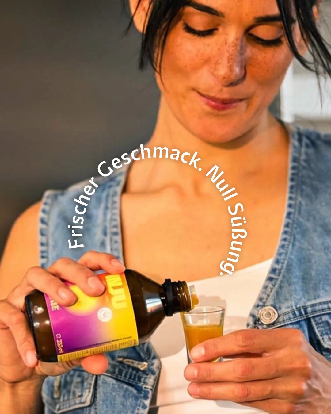Eine Frau schenkt sich INJU ONE aus der gelb-pinken Flasche in ein Shotglas ein. Text-Overlay: „Frischer Geschmack. Null Süßung.“