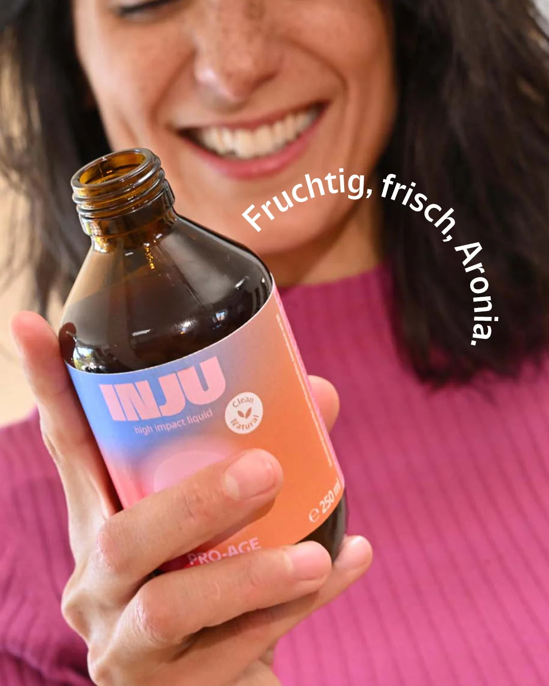 Eine lächelnde Frau hält die INJU Pro-Age Flasche. Text-Bogen: „Fruchtig, frisch, Aronia.“, was auf die Geschmacksrichtung der Aroniabeere hinweist.