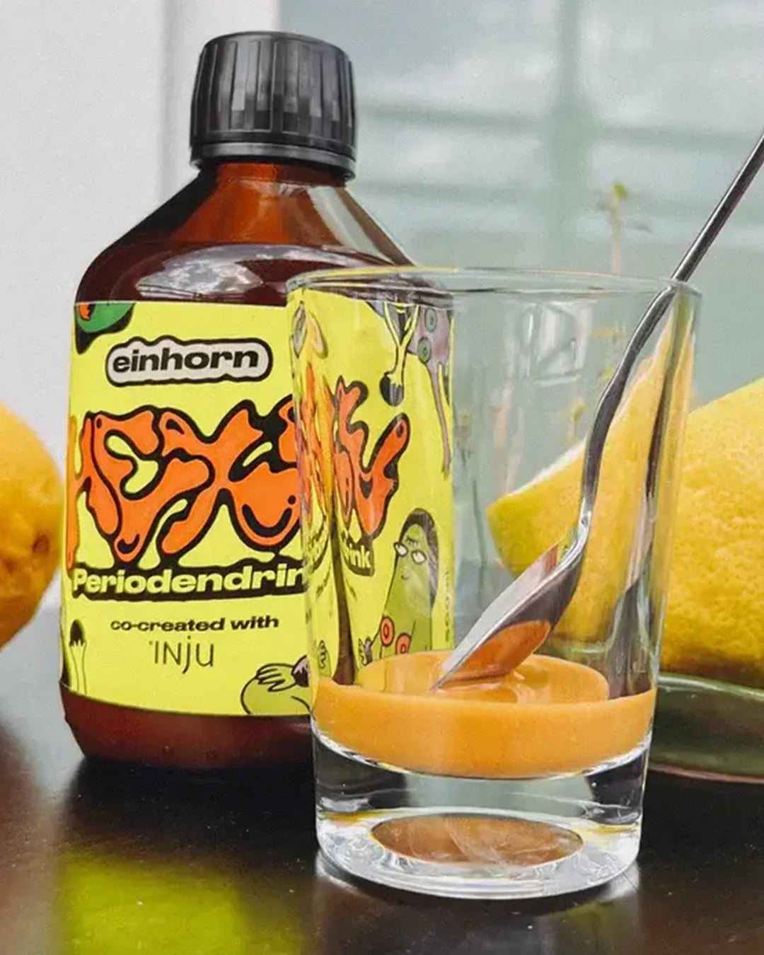 Eine braune Glasflasche des „Hexe Periodendrinks“ steht neben einem Glas, in dem sich die orangefarbene Essenz und ein Löffel befinden. Im Hintergrund sind frische Zitronen zu sehen.