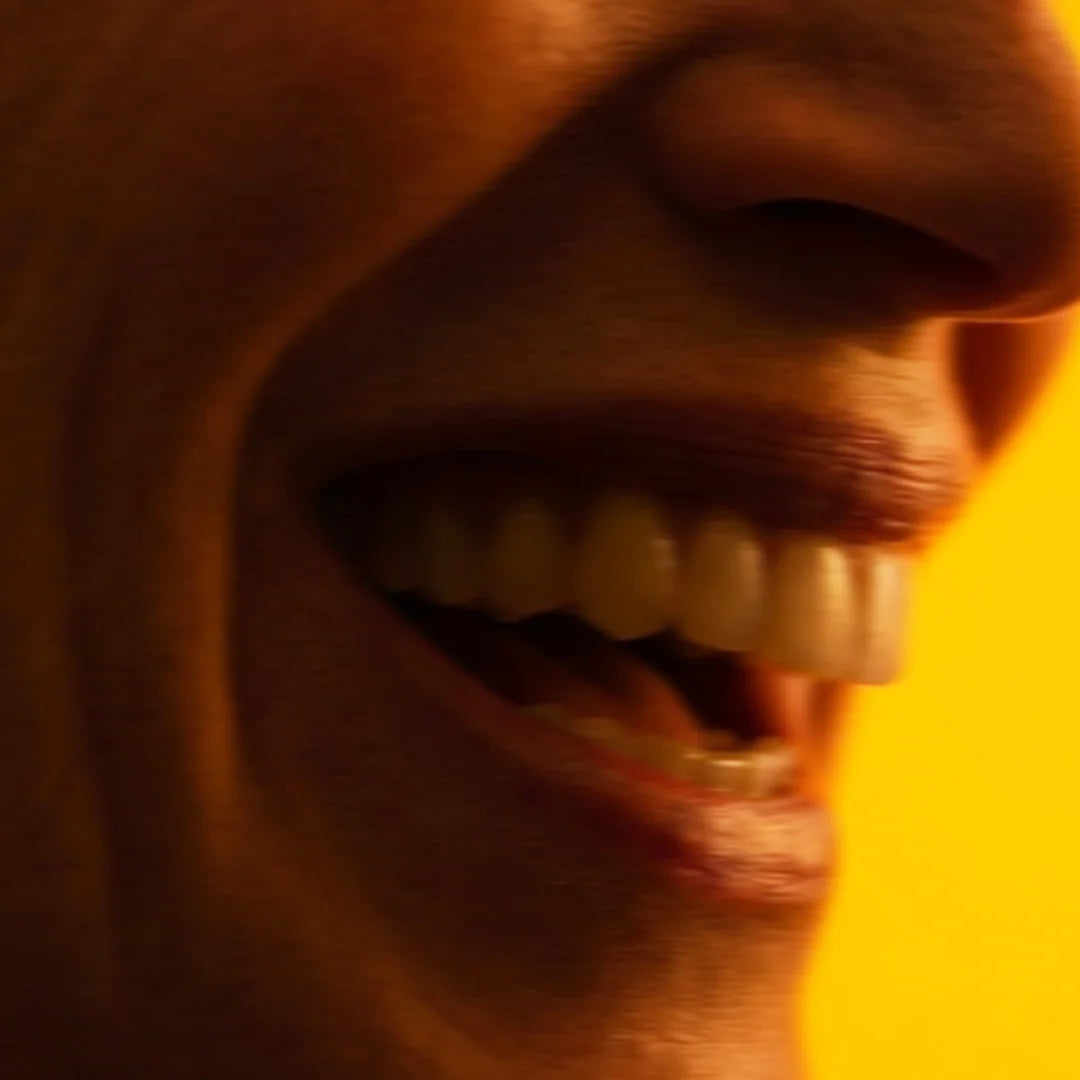 Nahaufnahme eines lachenden Mundes im Profil vor einem leuchtend gelb-orangefarbenen Hintergrund. Die oberen Zähne und die Lippen sind beim Lachen deutlich zu sehen, getaucht in warmes, atmosphärisches Licht.
